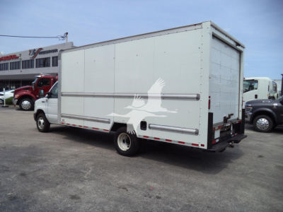 Ford E350 box truck rear three-quarter view with white cargo box, fuel tank, and dual rear doors; some text on distant building; stock/lot vehicles in background.