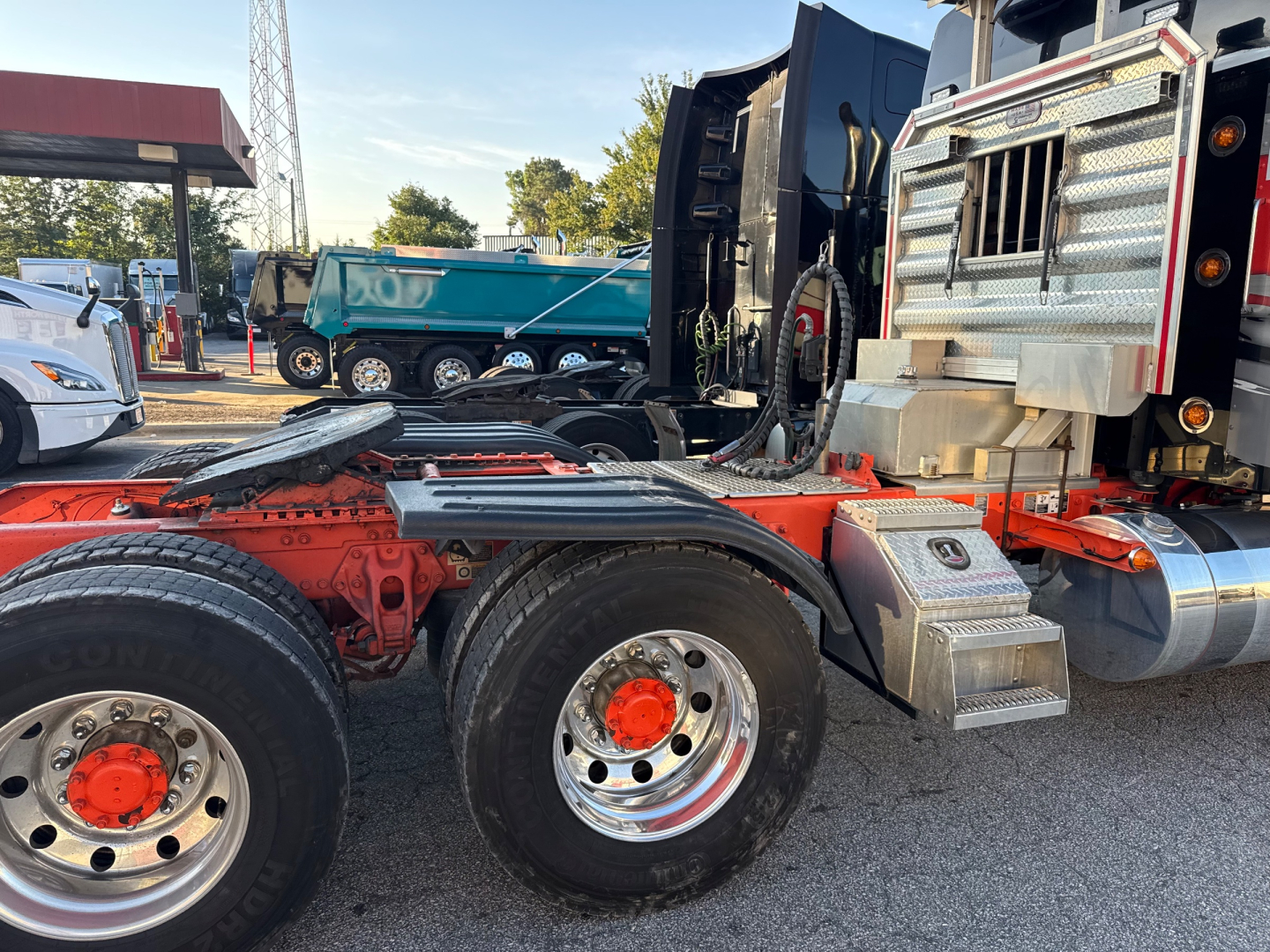Kenworth W900L truck chassis with orange frame and dual rear axles; metal diamond-plate toolbox; chrome fuel tank; black cab interior visible; background shows other trucks
