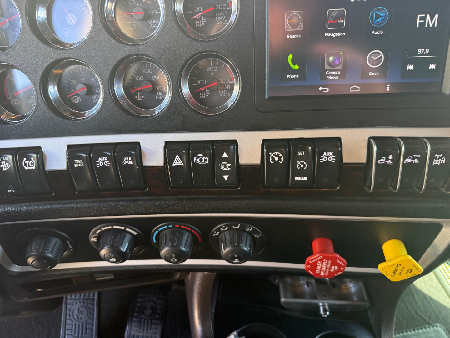 Cockpit of a 2022 Kenworth W900L showing multiple gauges, a touchscreen infotainment system, and various switches with red and yellow knobs.