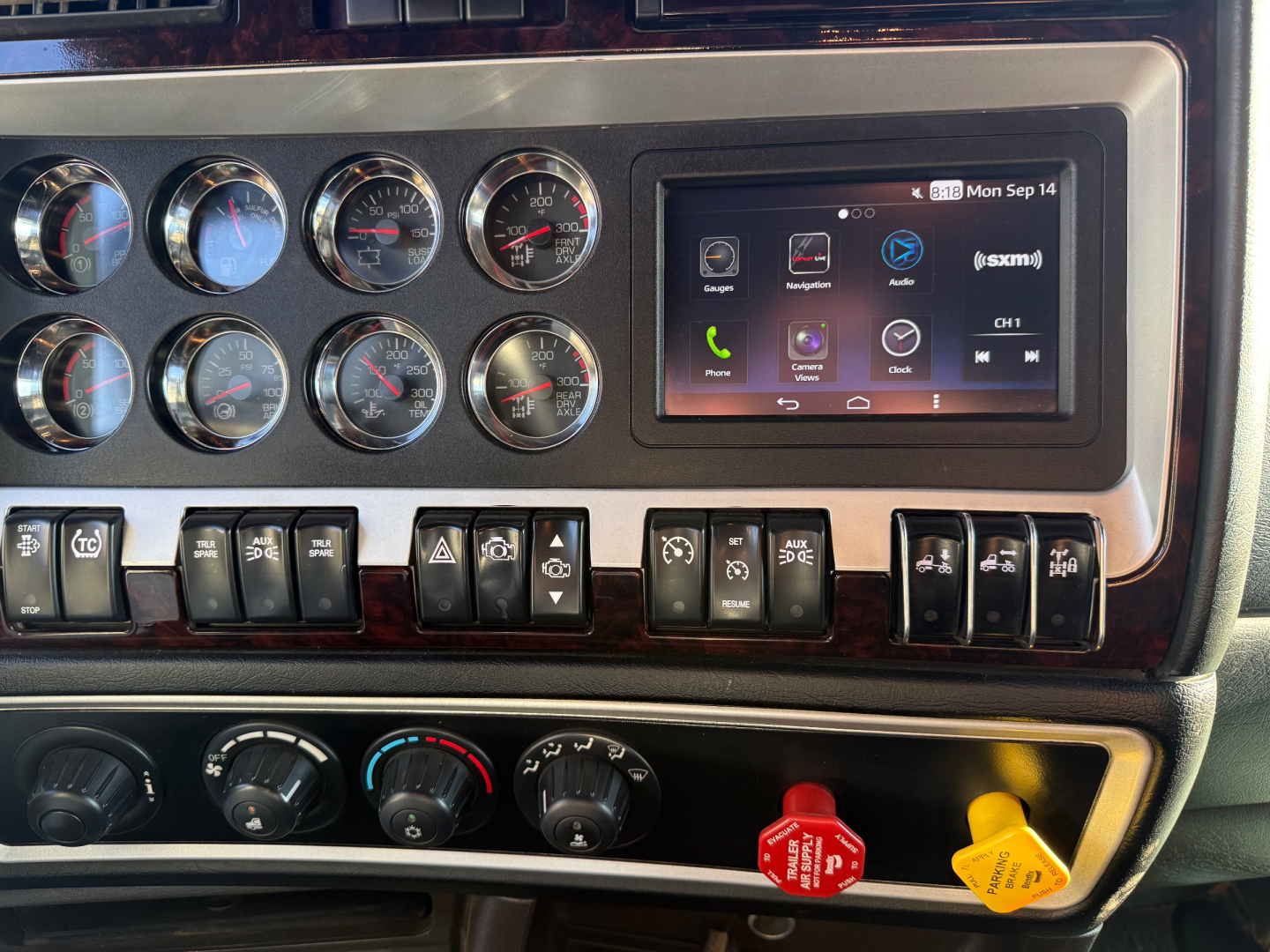 Kenworth W900L dash panel showing multiple round gauges, a touchscreen infotainment system, and various control buttons; visible knobs and a red emergency brake handle.