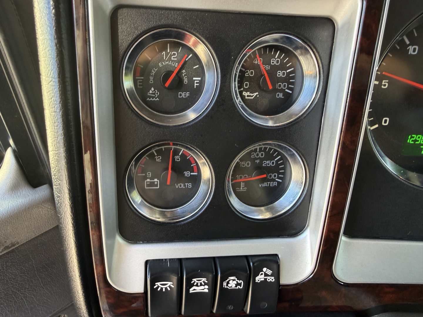 Kenworth W900L interior dashboard showing four gauges (E-DEF, volts, water temp) and switches located below, with steering column edge visible.