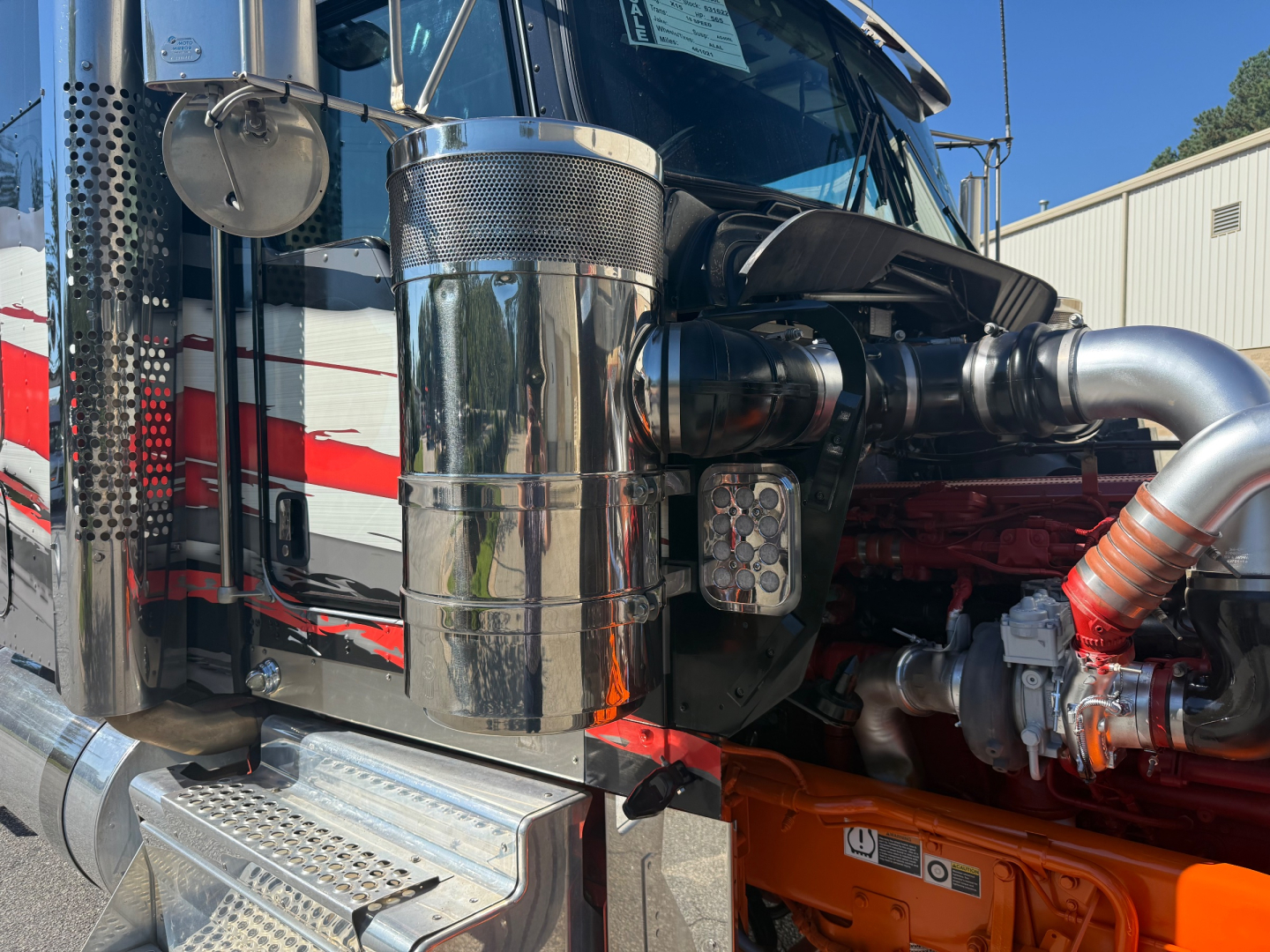 Kenworth W900L engine bay showing chrome air intake, coolant reservoir, turbocharger piping, and orange chassis frame.