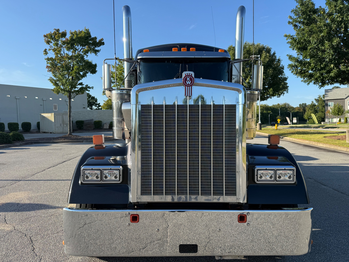 Kenworth W900L front view showing large chrome grille, dual vertical exhaust stacks, bumper with two red reflectors, headlights, and KW emblem on grill.