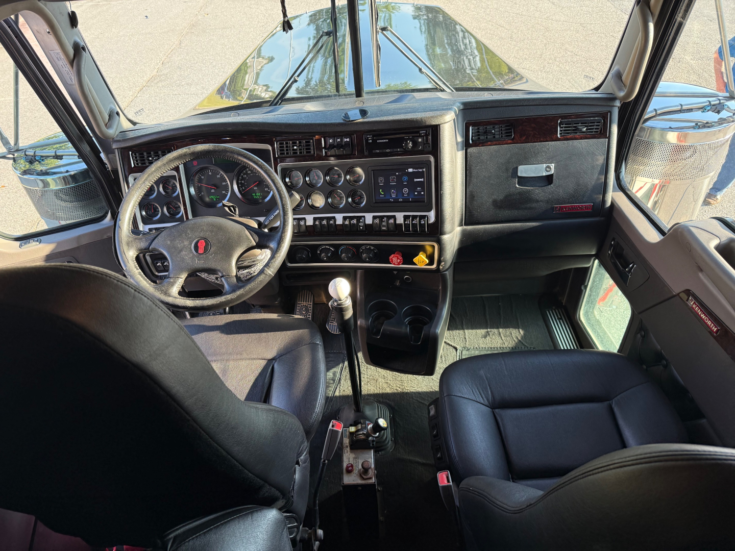 Kenworth W900L interior view showing steering wheel, dashboard with gauges, gear shifter, and driver seat in a conventional sleeper truck