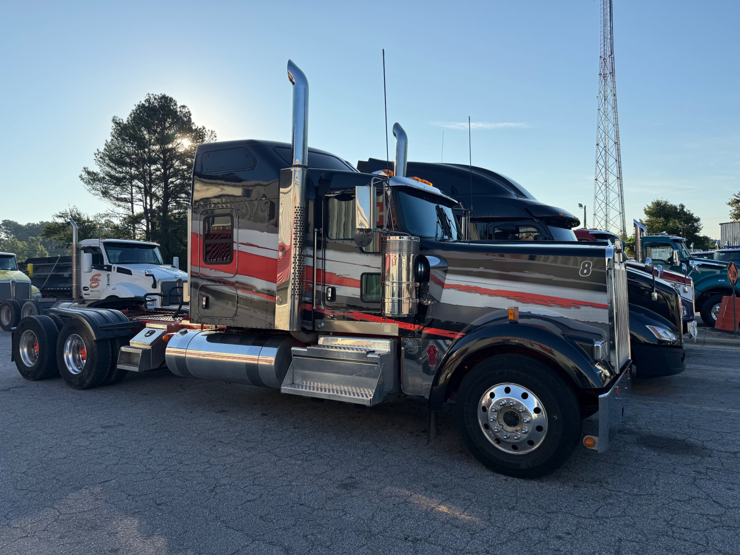 Kenworth W900L conventional sleeper truck shown in a lot, chrome exhaust stacks, side steps, and dual rear axles visible.