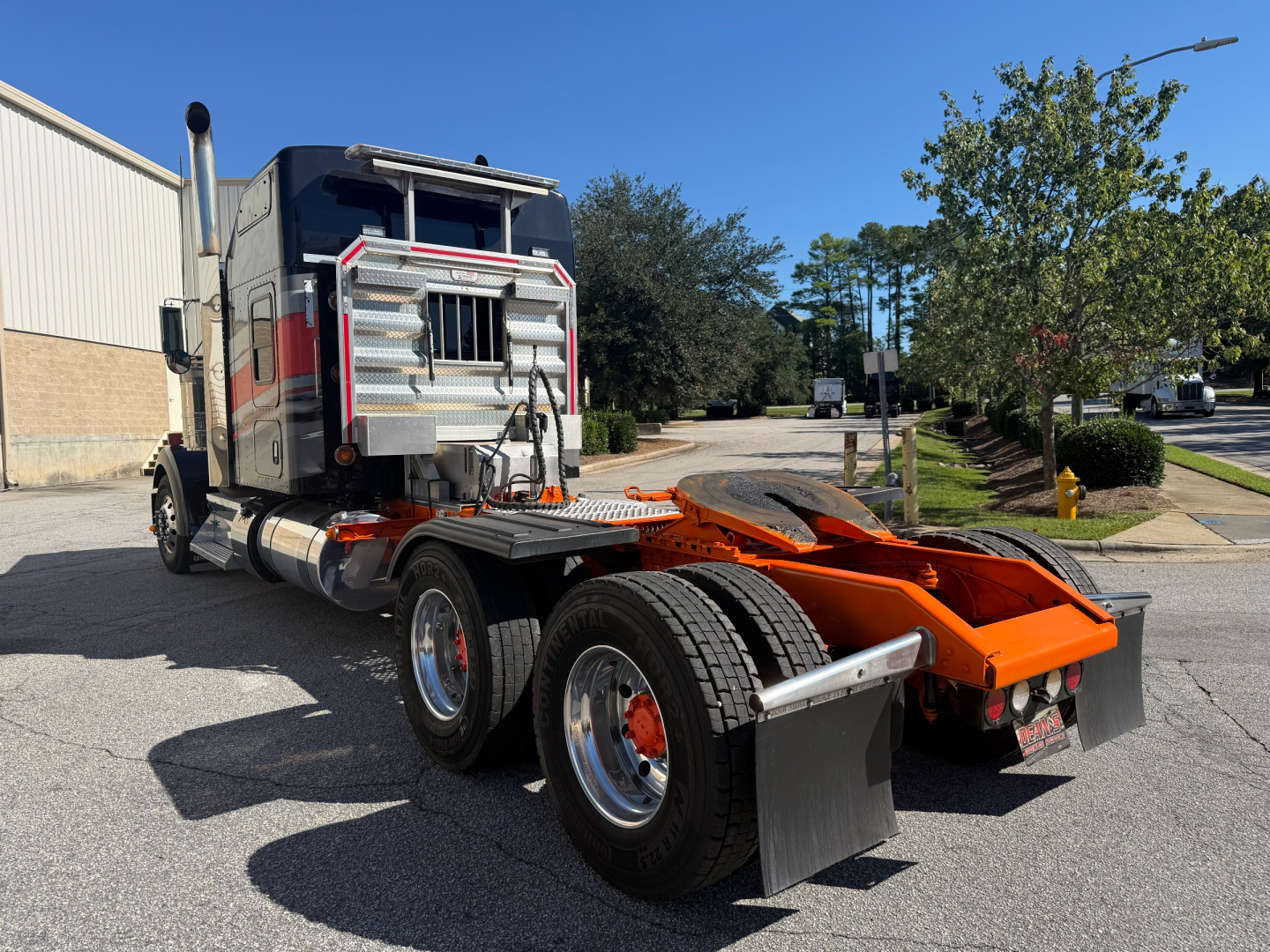 Kenworth W900L conventional sleeper truck rear view showing cab, dual rear axles, and orange fifth wheel/grabber in a parking lot.