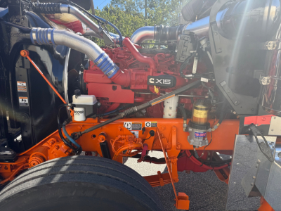 Kenworth W900L engine bay showing Cummins X15 engine and orange chassis components with hoses and reservoirs visible.