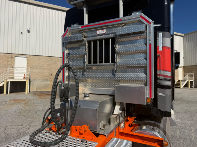 Kenworth W900L rear/side view showing aluminum diamond-plate toolbox, chrome exhaust stack trim, and bright orange hitch/step area.