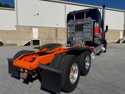 Kenworth W900L tractor with orange chassis and dual rear axles, cab interior visible, front grille and exhaust stack, aluminum fuel tanks, in a parking lot.