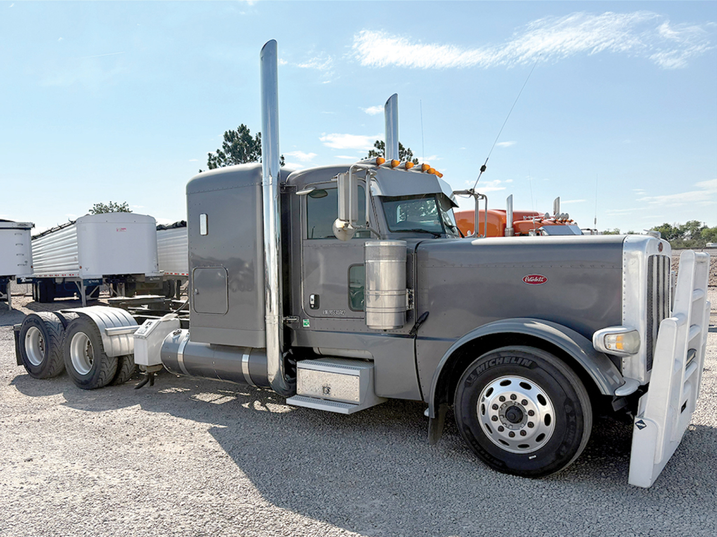 Peterbilt 389 extended hood semi truck side view with sleeper, gray color, chrome exhaust stacks, front bumper, and dual rear axles in a lot.