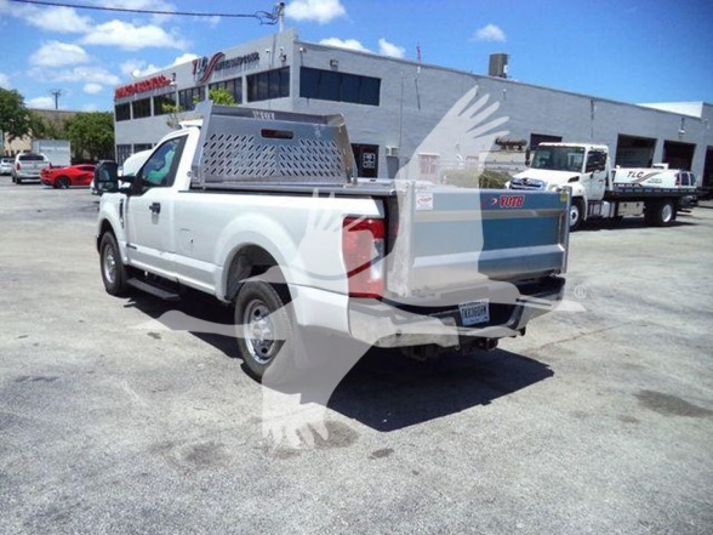 Ford F-250 pickup truck in a dealership lot, white utility dump bed with metal toolbox on bed and rear tailgate closed, rear three-quarter view with dealership building in background.