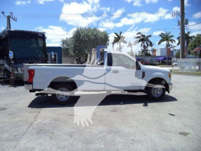 Ford F-250 pickup truck in a lot, side view, white with extended cab, long bed. Background shows palm trees, power lines, other vehicles.