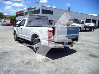 Ford F-250 pickup truck in a dealership lot, white utility dump bed with metal toolbox on bed and rear tailgate closed, rear three-quarter view with dealership building in background.
