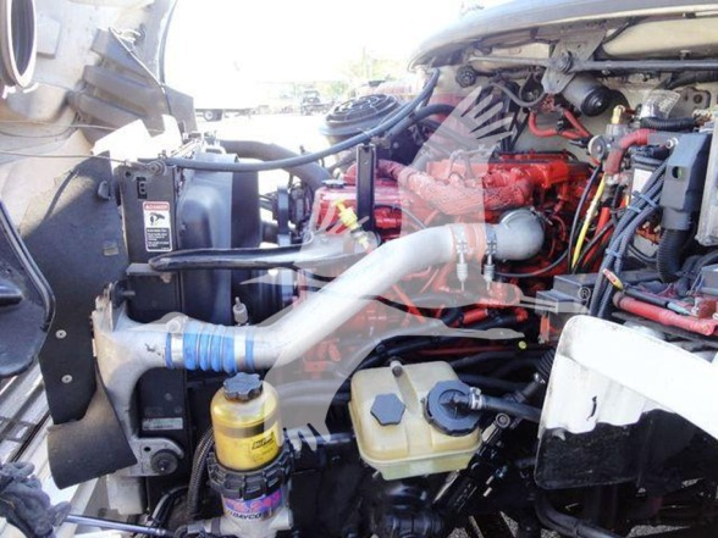 Front engine bay of a Freightliner Business Class M2 106 showing coolant reservoir, brake fluid reservoir, yellow oil filter area, red engine block, and various hoses.