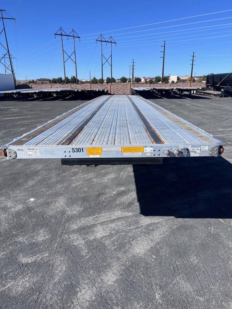 Flatbed trailer viewed from front center showing long aluminum deck with ribbed surface and wheel ramps at rear.