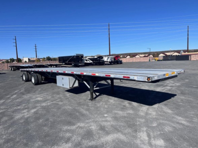 Flatbed trailer with dual rear axles, long gray deck, in a parking lot. Visible components include storage box under deck and support legs. No readable branding or model text visible on trailer.