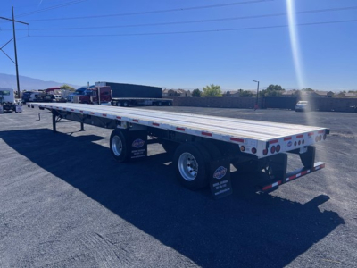 Flatbed trailer showing dual rear axles and long aluminum deck with reflective tape and rear lights.