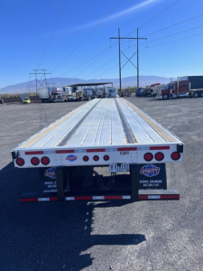 Flatbed trailer back view showing aluminum deck with multiple bright red circular taillights and Utility brand stickers; license plate area and rear bumper visible.
