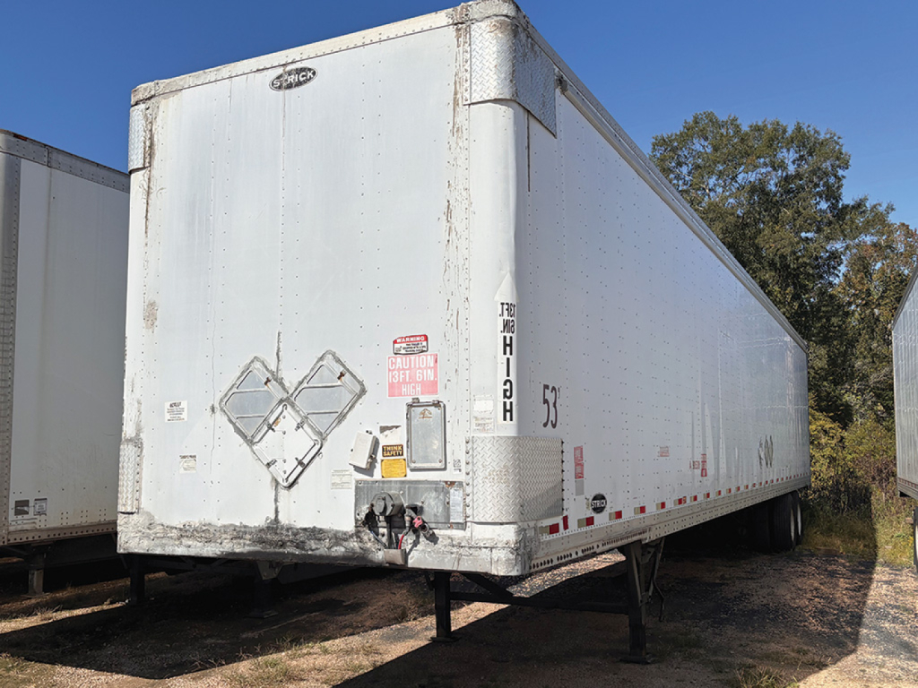 Rear three-quarter view of a white aluminum dry van trailer showing bumper and landing legs, with hazard placards and identification decals.