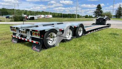 Trailer chassis with multi-axle lowboy trailer showing steel deck, dual rear axles, and rear lights in a grassy lot.