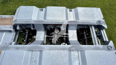 Trailer deck with exposed mechanical components and wiring in a lowboy trailer, showing metal cross members and engine-like assemblies under a grated aluminum surface.