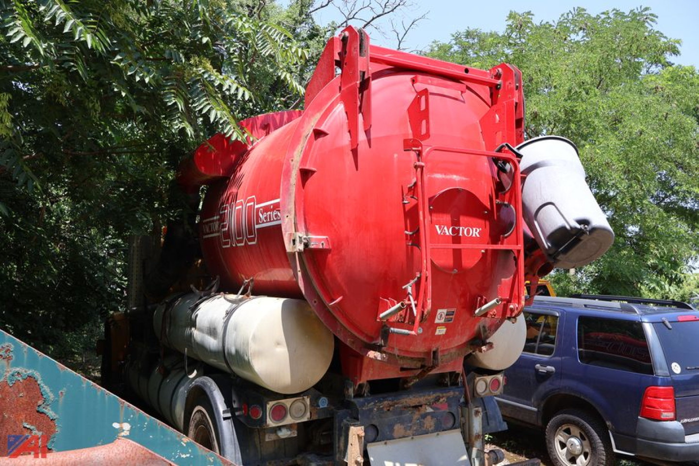 Vactor vacuum tank truck rear view showing red cylindrical tank with hoses and white tank on side, International 4900 branding faint, truck bed and green trees in background.