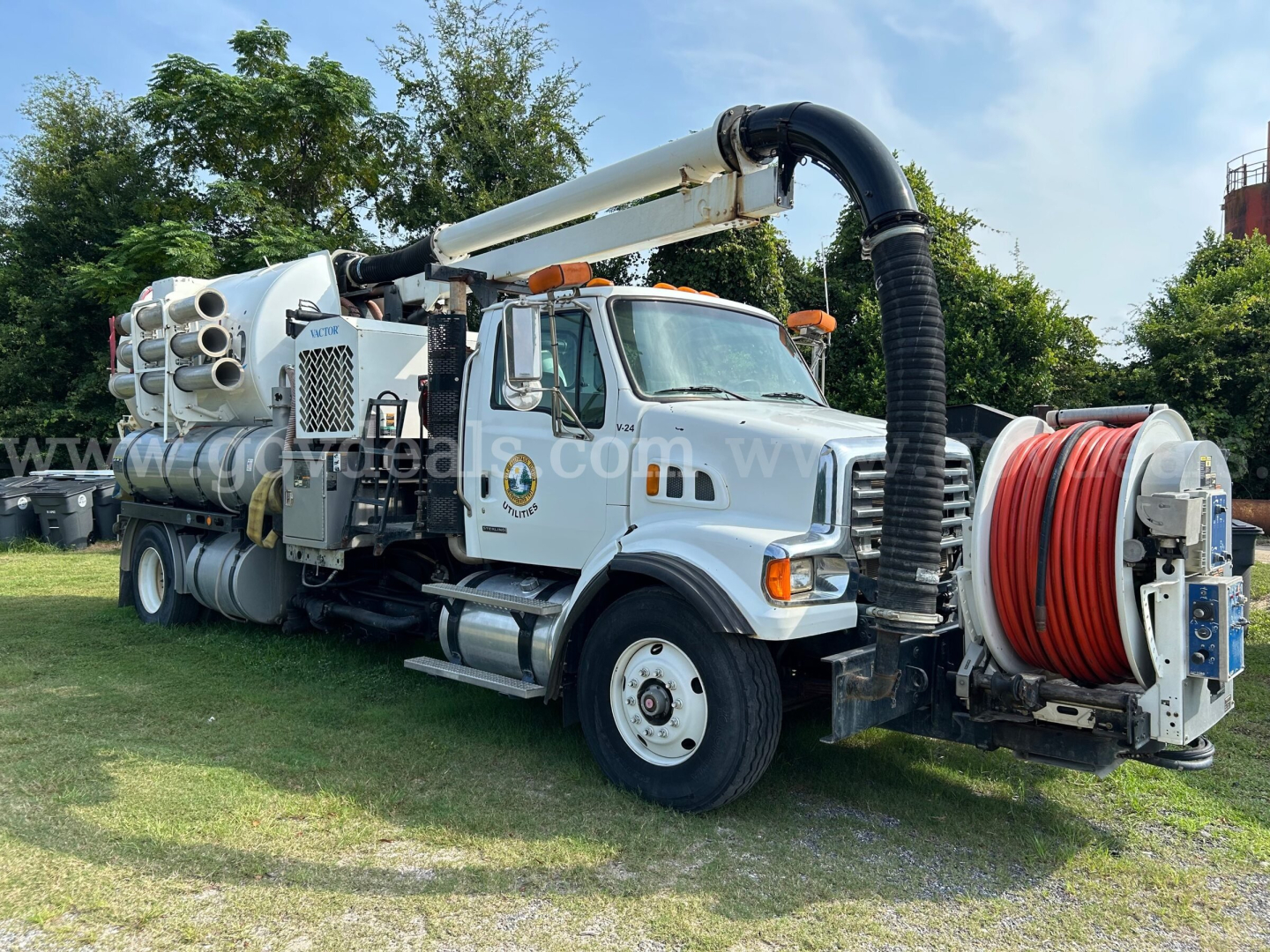 Concrete pump truck with long articulating boom and hose reel visible on side, silver cab, large black suction hose curling over top, red hose reel at front, multiple cylindrical tanks along chassis.