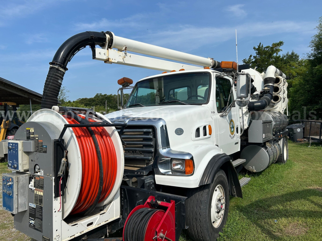 Truck chassis with large vacuum hose reel and hose reels; front grille and cab visible; high pressure vacuum system mounted on back; white truck with black hose, red and white hose reels, and hydraulic arm.