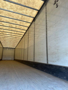 Trailer interior showing long empty cargo space with wooden floor and white side walls, top translucent roof panels, and metal support beams.