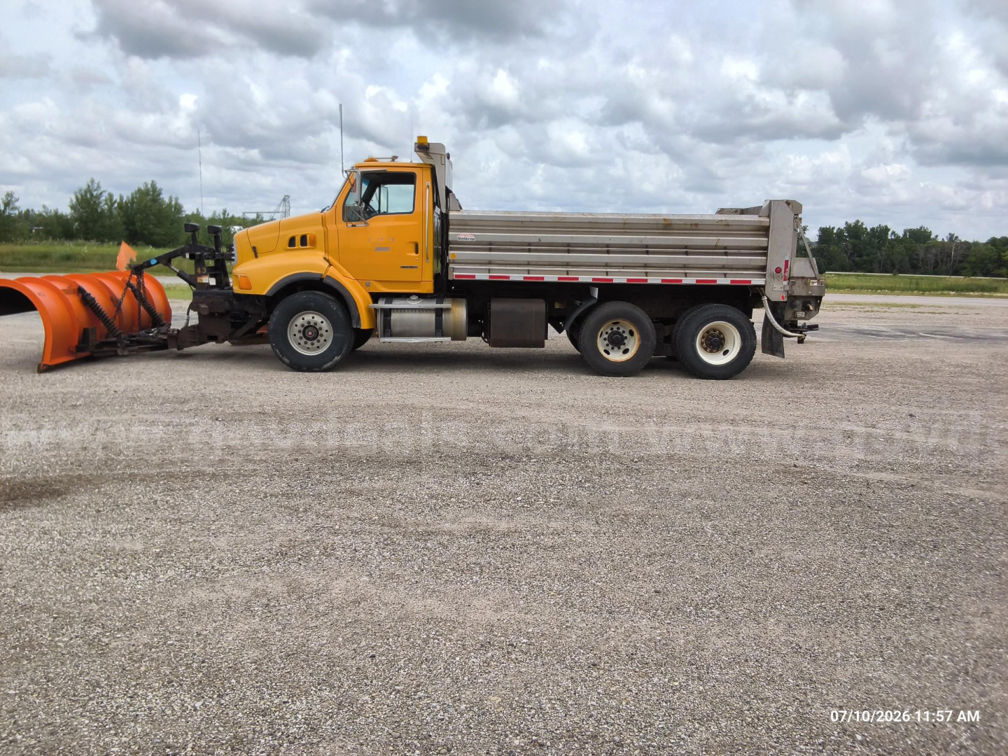 Dump truck attached to snow plow blade, yellow cab and gray dump bed, side view, on gravel lot.