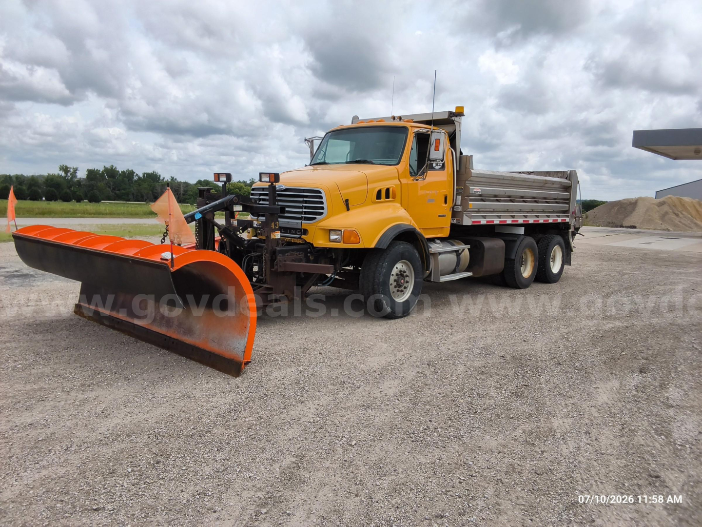 Dump truck with a bright yellow Sterling LT8500 cab and a large orange snow plow attachment in front, dumping bed visible in rear, on gravel lot under cloudy sky.