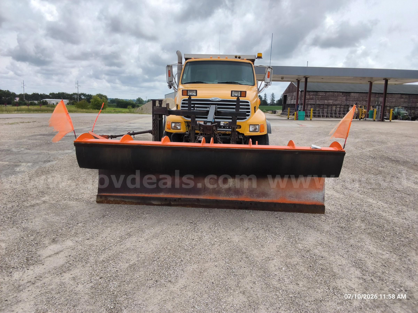 Front view of a yellow Sterling LT8500 dump truck with a large black snow plow attachment and orange safety flags.