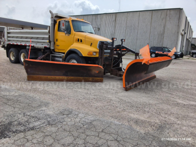 Heavy-duty dump truck with a wide snowplow blade attached, yellow cab, long dual rear wheels, and a large orange snow blade in front, warehouse in background.