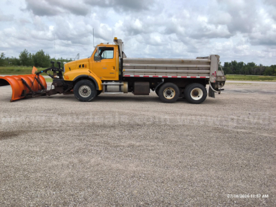 Dump truck attached to snow plow blade, yellow cab and gray dump bed, side view, on gravel lot.