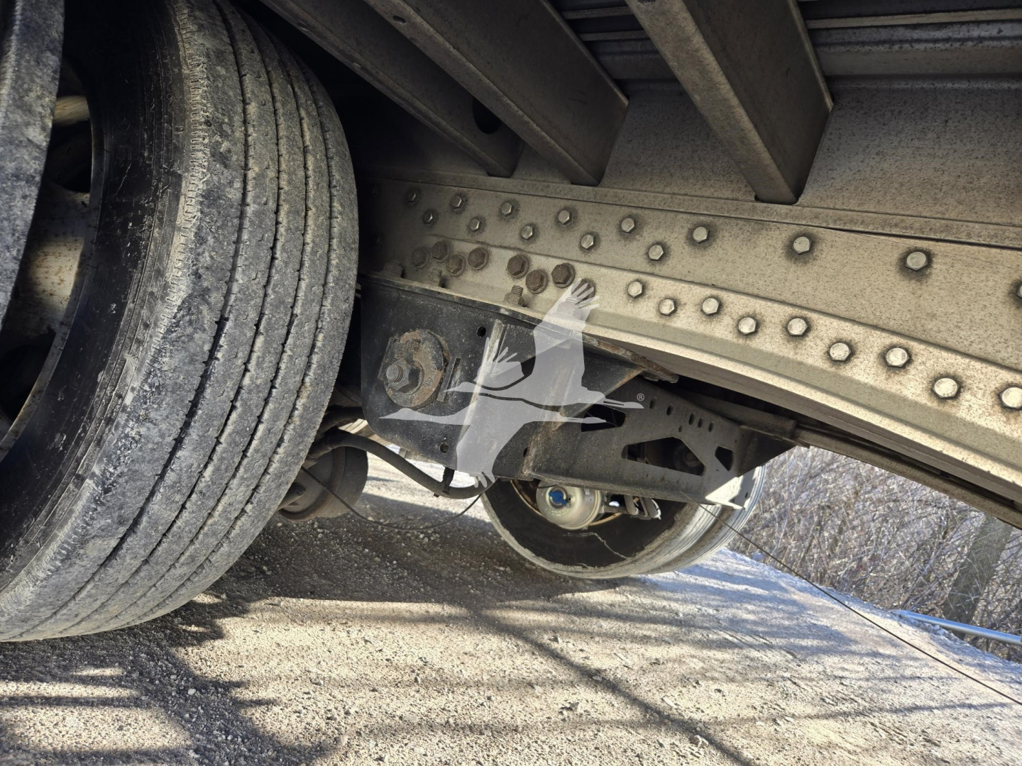 Underbody of a trailer showing riveted frame, leaf spring suspension, and dual tires. Visible components include a hinge/bolt assembly and undercarriage with metal beams.