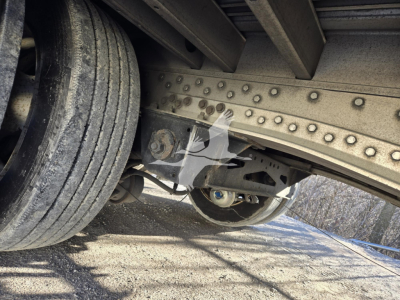 Underbody of a trailer showing riveted frame, leaf spring suspension, and dual tires. Visible components include a hinge/bolt assembly and undercarriage with metal beams.