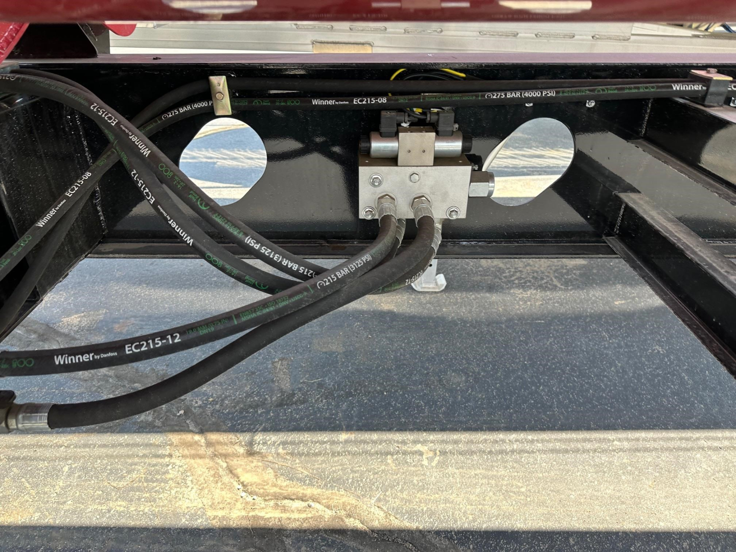 Trailer hitch area showing hydraulic lines and control valve mounted on a black frame with two circular cutouts and metal brackets.