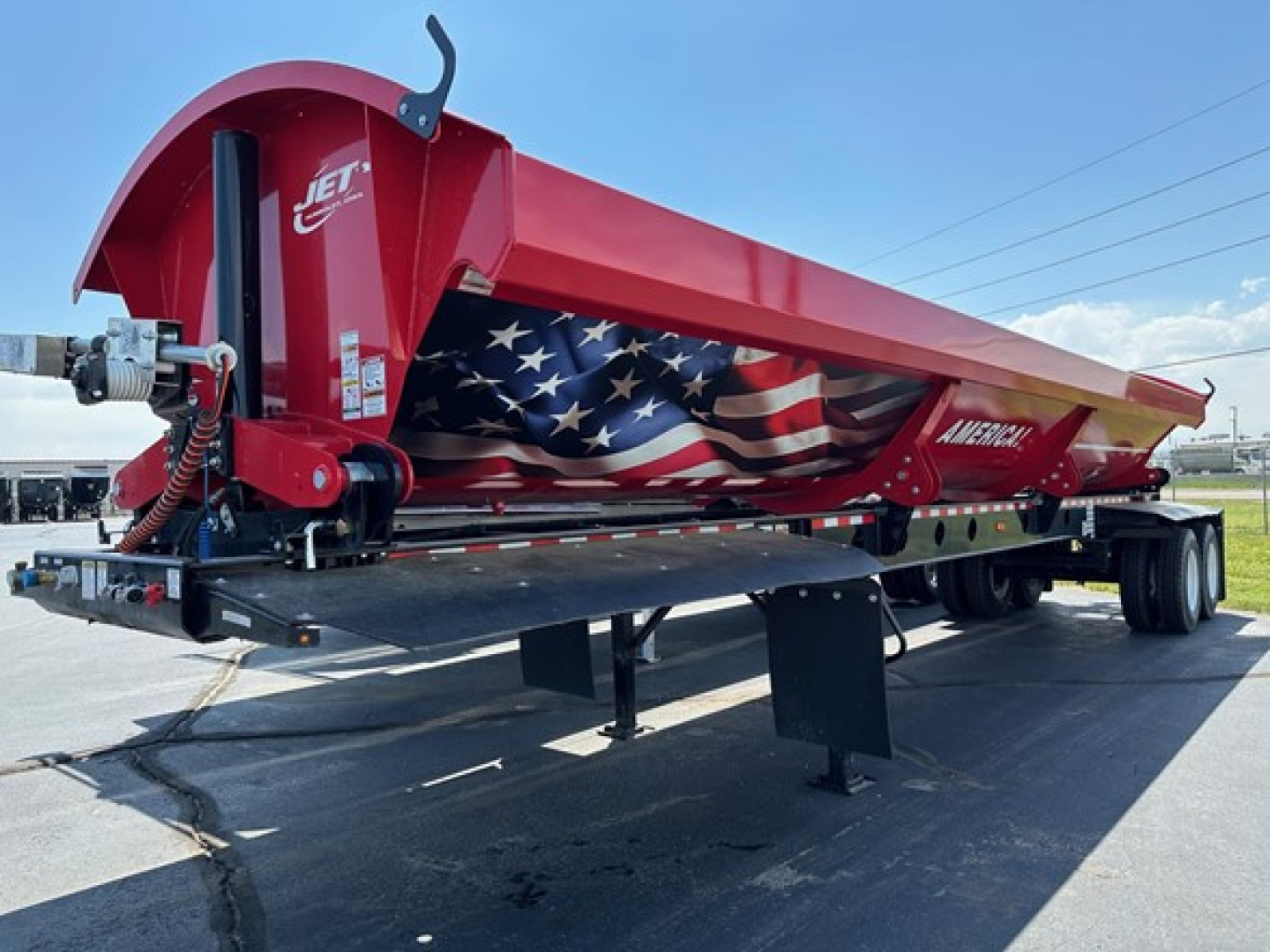 Jet side-dump trailer in red with American flag graphic along side, rear hitch area visible, landing gear and dump mechanism present, parked on pavement.