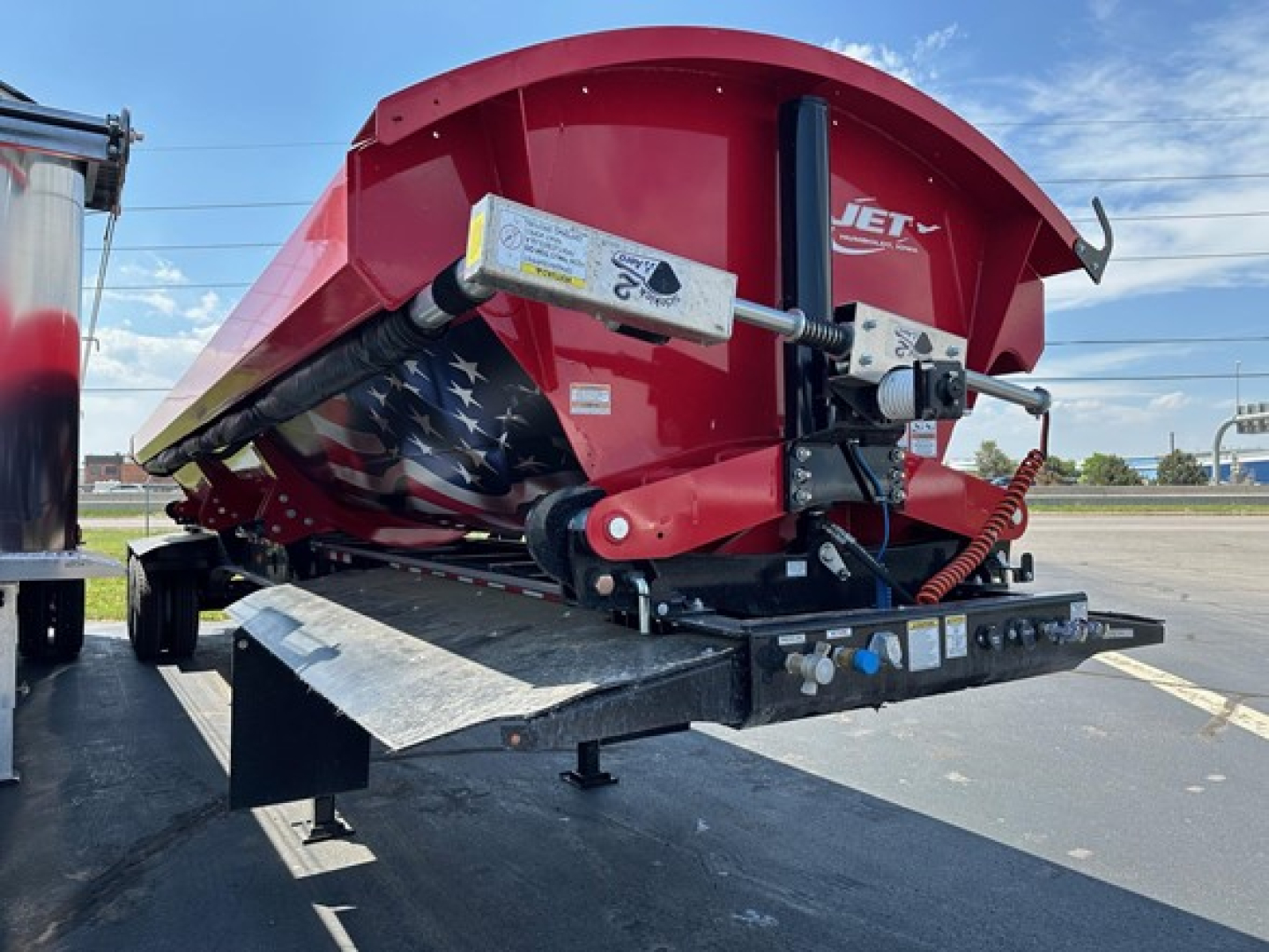 Jet Jet side dump trailer in red and black, on a flatbed, showing hydraulic cylinders, mounting hardware, and a chevron-like star-spangled pattern on the inner compartment.