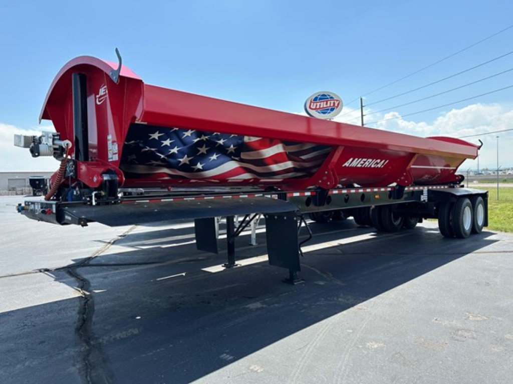 Red Jet side-dump trailer showing American flag graphics and Utility logo, parked on lot with triple-axle rear; model name Ameri on side.