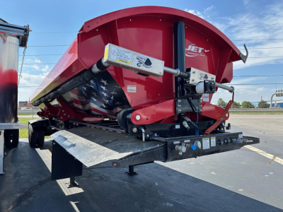 Jet Jet side dump trailer in red with hydraulic lift and metal framework, parked on trailer bed; visible safety decals on hydraulic cylinder and control box.
