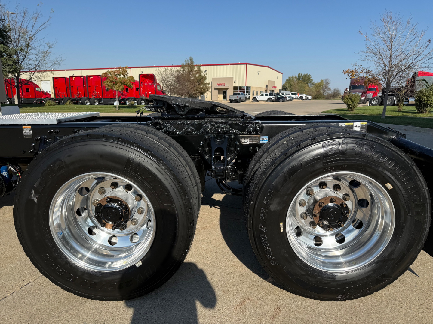 Peterbilt 579 dual rear wheels (tractor) with chrome wheels and frame, showing tires and fifth wheel area; background shows a truck dealership lot with red trucks and a beige building.