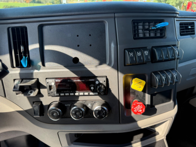 Truck dashboard panel of a 2023 Peterbilt 579 showing a radio head unit, climate control knobs, and various toggle switches; several colored caps (yellow, red) on the right side, and a blue tag hanging near the air vent.
