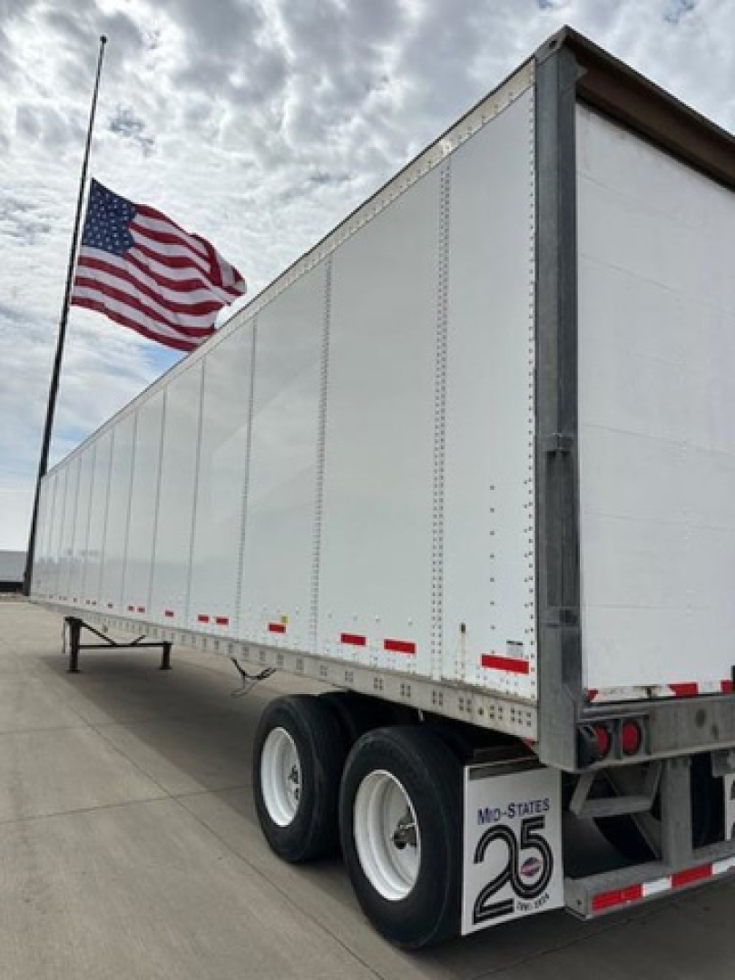 Trailer side and rear showing a white Wabash trailer with dual rear wheels and red reflective tape. American flag in background.