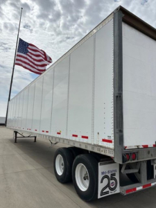 Trailer side and rear showing a white Wabash trailer with dual rear wheels and red reflective tape. American flag in background.