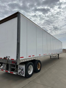 Trailer showing a long white Wabash van trailer with dual rear axles and reflective safety markings. Visible text includes license plate area and small stickers on wheels.