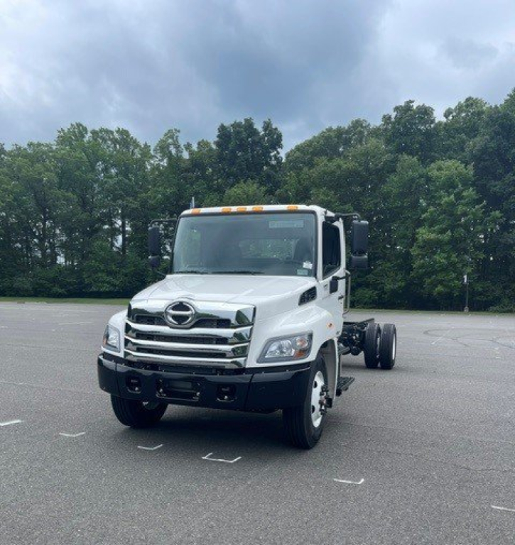 White 2027 Hino L7 cab chassis truck in a parking lot, front three-quarter view showing grille, headlights, and dual rear wheels not yet attached.