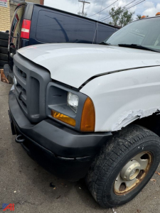 Front left view of a white 2005 Ford F350 XL SD pickup truck with damaged front fender and exposed wheel, black bumper, amber turn signal, and a blue van in the background.