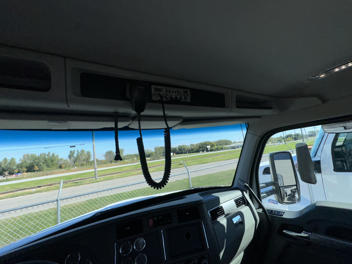 Kenworth T880 cab interior with overhead dashboard and CB microphone; windshield and side mirror visible; exterior road and fence in background.