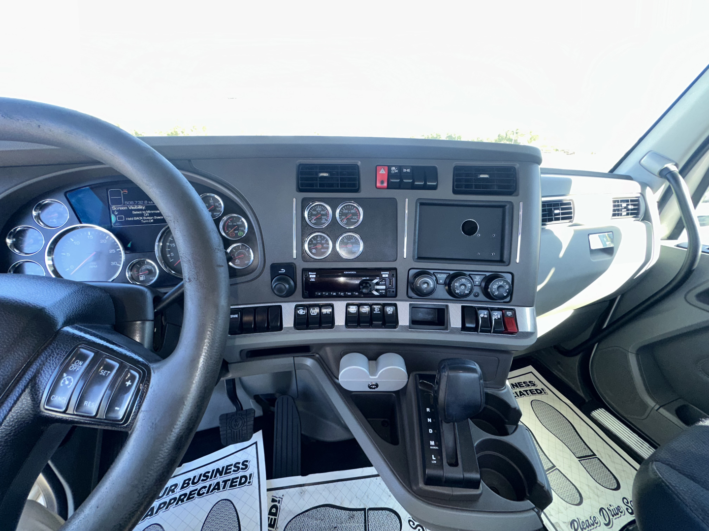 Kenworth truck interior dashboard with multiple gauges, steering wheel, gear shifter, and center console gauges. Text visible includes various gauges and controls but no legible model details beyond general dashboard components.